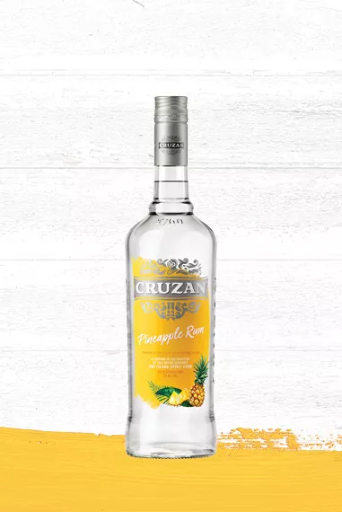 Clear glass bottle of Cruzan Pineapple Rum with a yellow label set against a whitewashed wood background with a pineapple yellow paint swash.