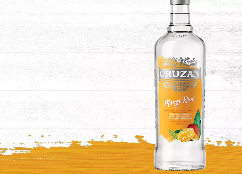 Glass Cruzan Mango Rum bottle against a whitewashed wood background with a mango-hued paint swash.