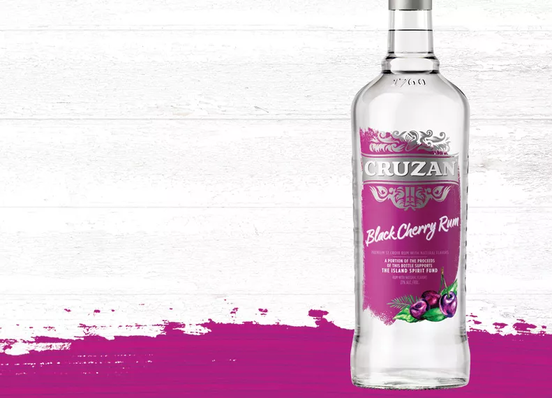 Glass bottle of Cruzan Black Cherry Rum with a magenta label decorated with black cherries set against a whitewashed wood background with a magenta paint swash.