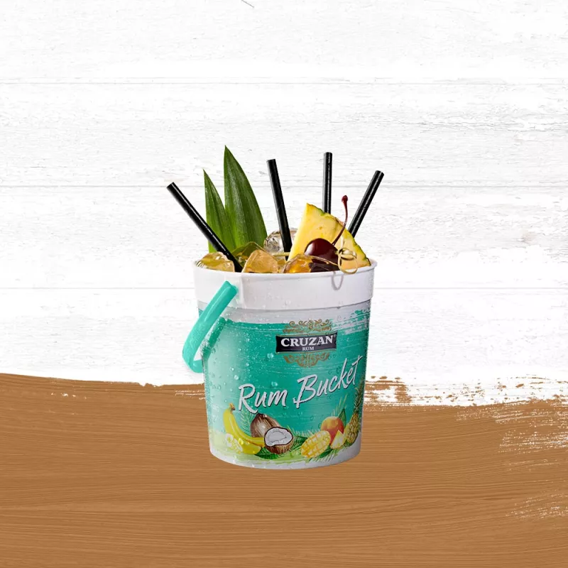 Rum Runner cocktail in a Cruzan® Rum bucket with drinking straws and garnished with a pineapple wedge and cherry set against a whitewashed wood background with a gold paint swash.