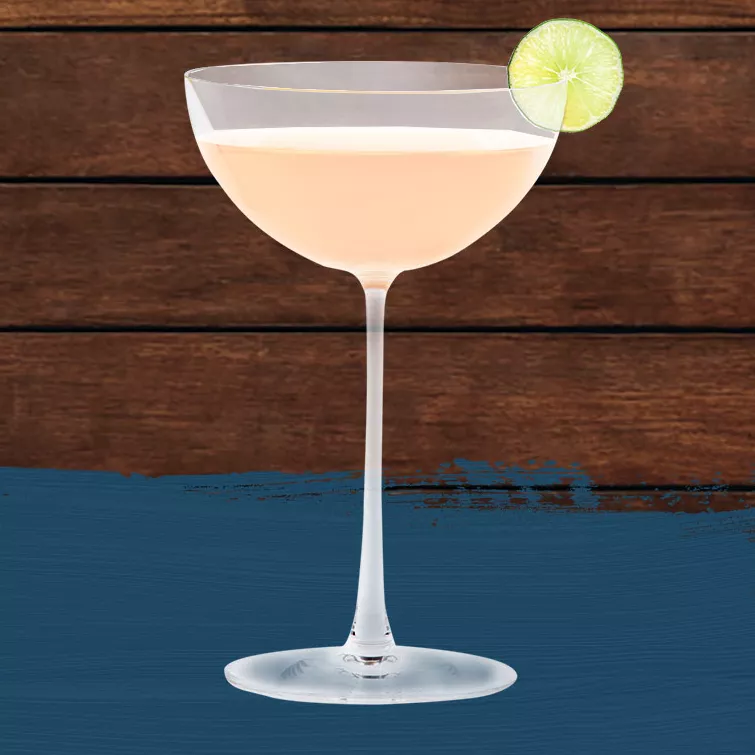 Hemingway Daiquiri in a coupe glass, garnished with a lime wheel set against a wood background with a dark blue paint swash.