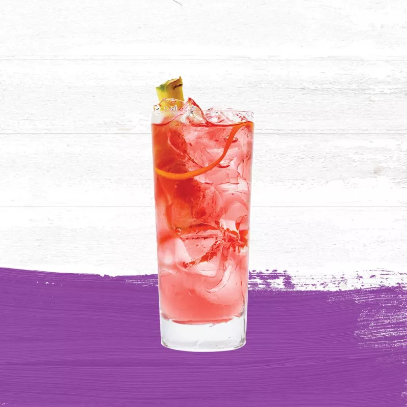 Passion Punch in a tall glass, garnished with pineapple and an orange rind set against a whitewashed wood background with a purple paint swash.