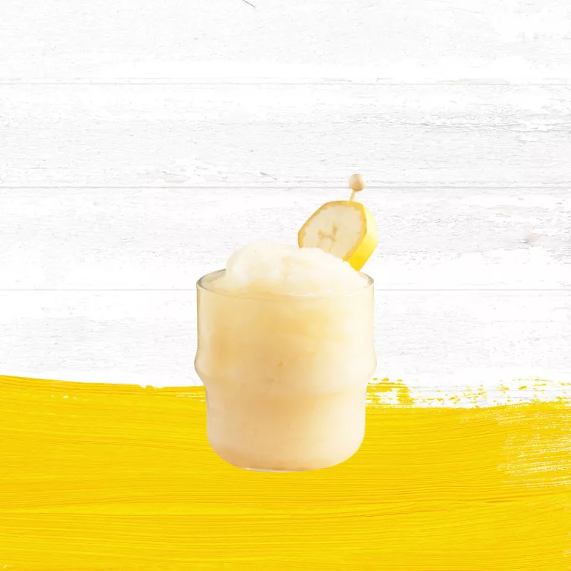 Banana Nana cocktail in a clear rocks glass garnished with a banana slice set against a whitewashed wood background with a yellow paint swash.