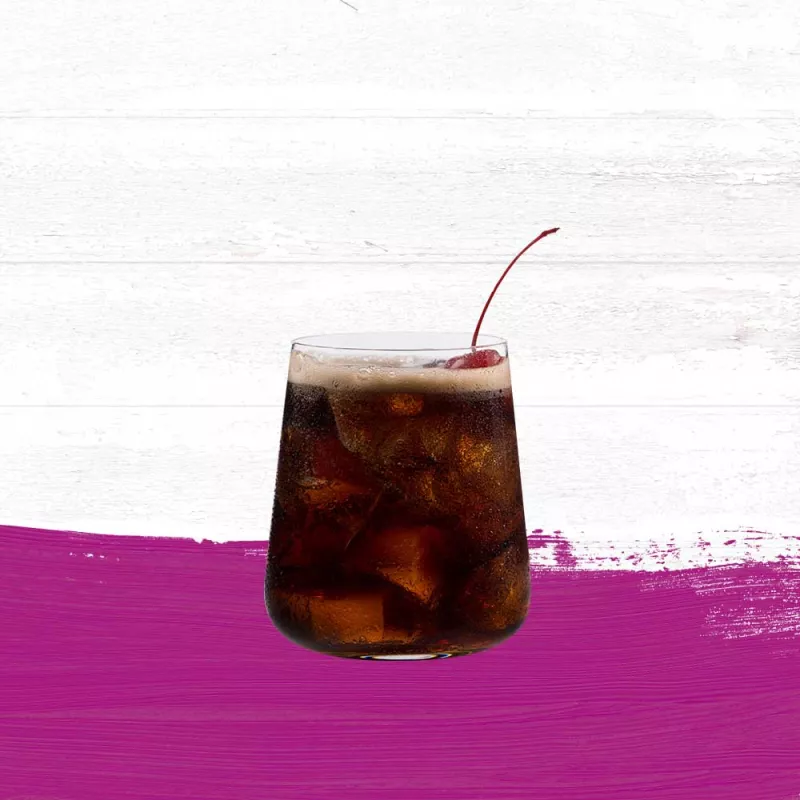 Black Cherry Rum & Cola in a small cocktail glass garnished with a cherry set against a whitewashed wood background with a cherry red paint swash.