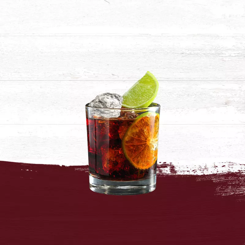Rum & Cola cocktail in a clear glass, garnished with a slice of lime against a whitewashed wood background and deep red paint swash.