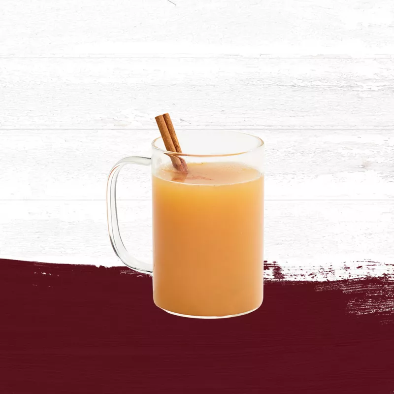 Hot apple cider mixed with Cruzan® Island Spiced Rum in a clear mug garnished with a cinnamon stick set against a whitewashed wood background with a deep red paint swash.