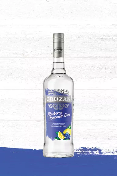 Set against a whitewashed wood background is a clear glass bottle of Cruzan Blueberry Lemonade Rum with a blue label and decorated with blueberries and lemon wedges.