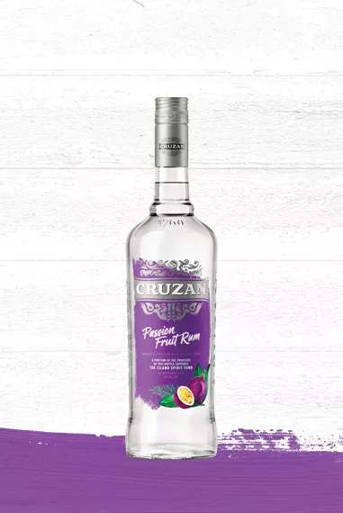 Set against a whitewashed wood background with a purple paint swash is a clear glass bottle of Cruzan Passion Fruit Rum with a royal purple label and adorned with passion fruit.
