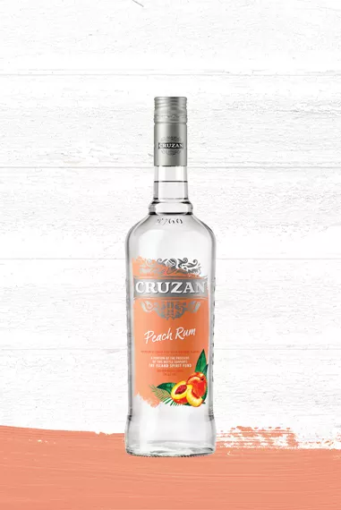 Clear glass bottle of Cruzan Peach Rum with a peach label set against a whitewashed wood background with a peach colored paint swash.