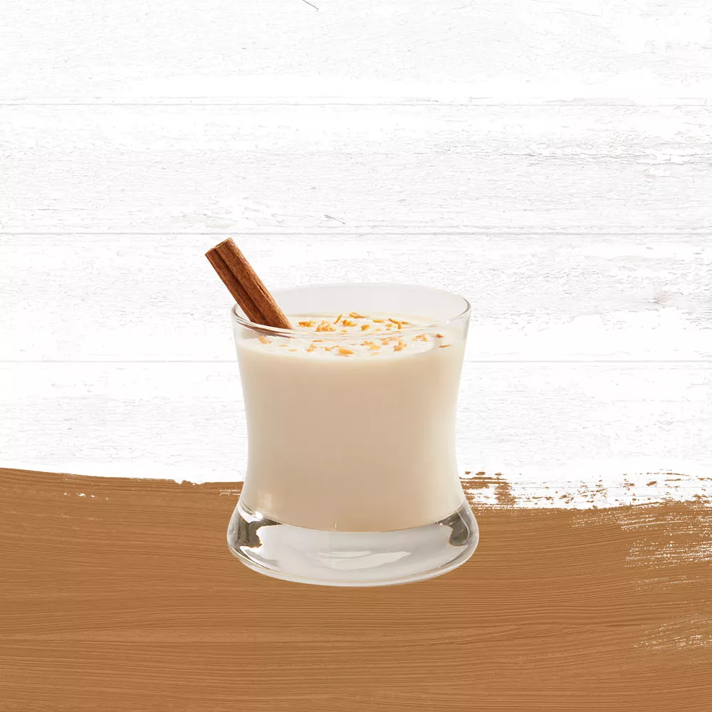 Coquito cocktail garnished with a cinnamon stick and shredded coconut in a rocks glass against a whitewashed wood background with a gold paint swash.