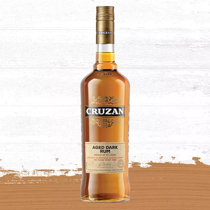 Glass bottle of Cruzan Aged Dark Rum against a background of light oak barrel panels and a light wood-colored paint smudge.