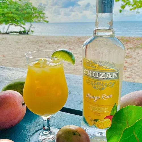 View of the Cruzan Mango Rum bottle with a mixed drink on the beach.