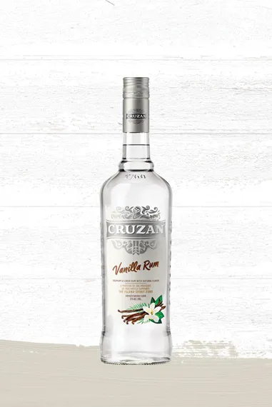 Set against a whitewashed wood background is a clear glass bottle of Cruzan Vanilla Rum with a white label adorned with vanilla pods and flower.